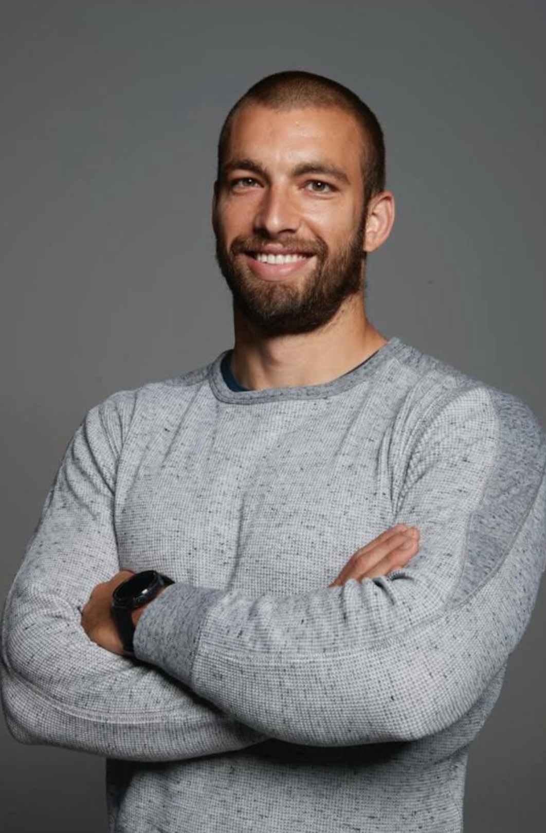 Uomo con barba corta e capelli rasati indossa un maglione grigio a trama fine. Ha le braccia incrociate e un orologio nero al polso, con uno sfondo neutro. Espressione sorridente e amichevole.